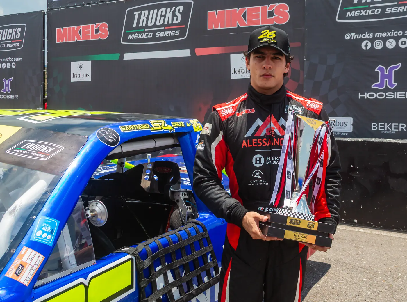 Santiago Cruz con su traje de carreras y un trofeo, junto a su camioneta, frente a la lona de Trucks México Series.